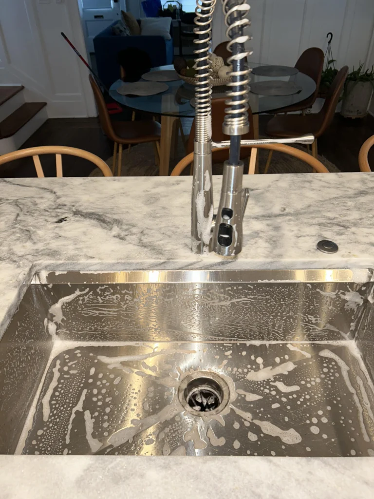 Stainless steel kitchen sink and pull-down faucet being scrubbed with foaming cleaner during a detailed deep-cleaning service.