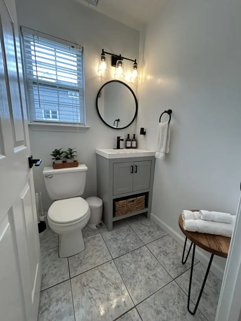 Bright, freshly cleaned bathroom with gray vanity, round mirror, white towels, polished tile floors, and neatly arranged decor for a welcoming guest-ready finish.