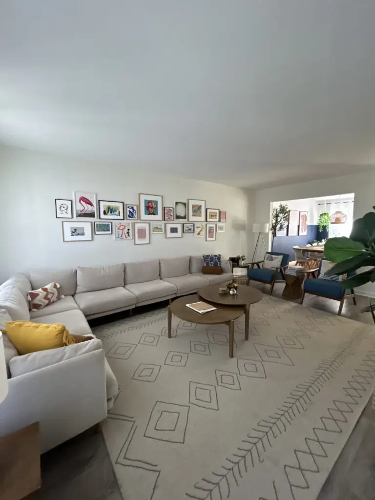 Bright living room with a large cream sectional, patterned area rug, round coffee table, colorful gallery wall, accent chairs, plants, and an open view into the dining area.