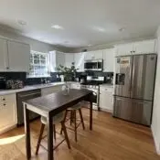 Bright kitchen with white cabinets, stainless steel appliances, black tile backsplash, polished hardwood floors, and a neatly cleaned island table with stools and greenery.