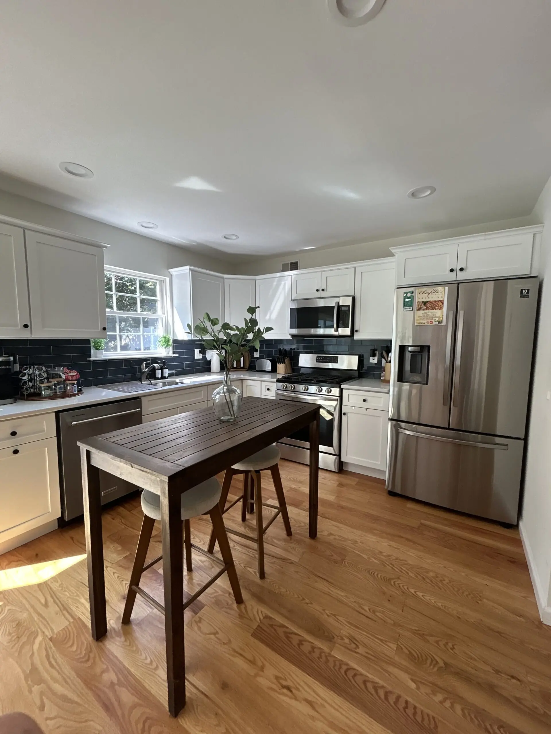 Bright kitchen with white cabinets, stainless steel appliances, black tile backsplash, polished hardwood floors, and a neatly cleaned island table with stools and greenery.