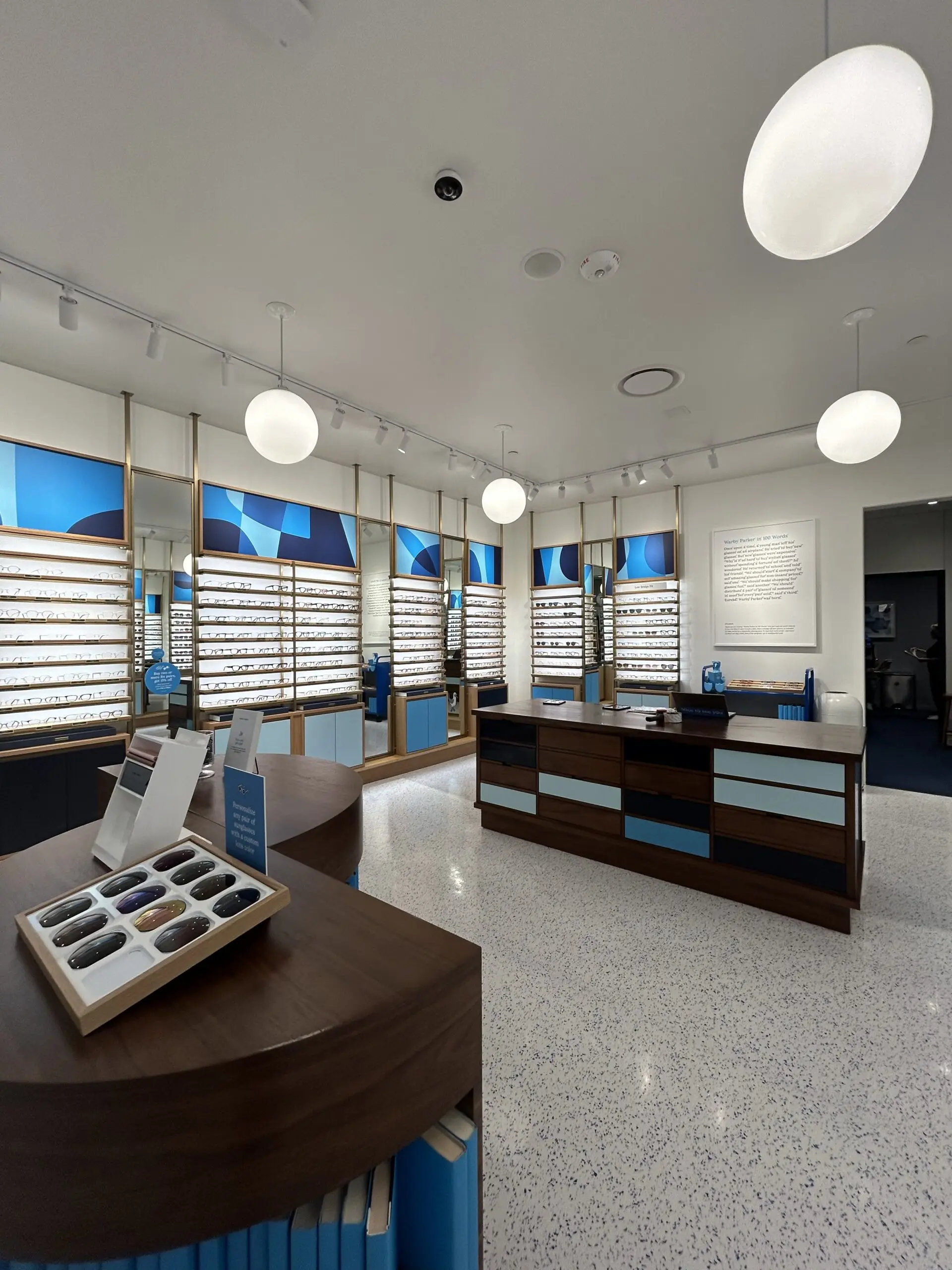 Bright optical retail store with neatly arranged eyeglass displays, polished speckled floors, modern globe lighting, blue accent décor, and organized service counters.