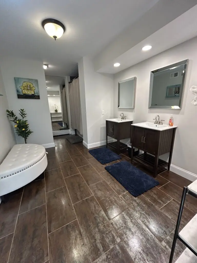 Clean, spacious bathroom with double vanities, polished mirrors, fresh rugs, shiny tile flooring, seating area, and a neatly maintained shower space after professional cleaning.