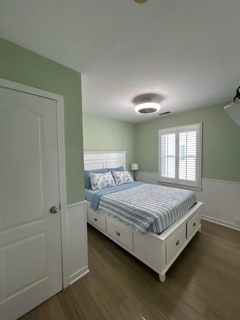 Clean and neatly prepared bedroom with a made bed, fresh linens, polished floors, white furniture, soft green walls, and natural light coming through the window shutters.