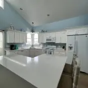 Clean, bright kitchen with white cabinets, polished countertops, white appliances, blue walls, vaulted ceiling, and a spacious island after professional cleaning.