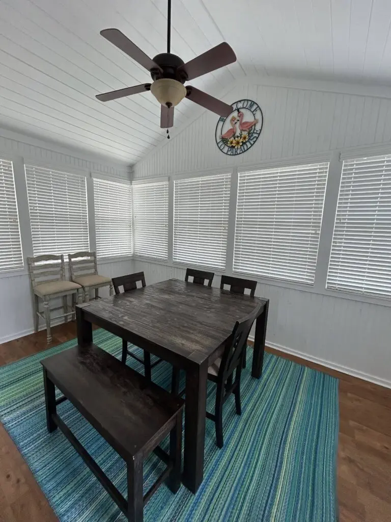Clean coastal dining room with a dark wood table, bench seating, bright window blinds, white paneled walls, ceiling fan, and a striped blue-green rug after professional cleaning.