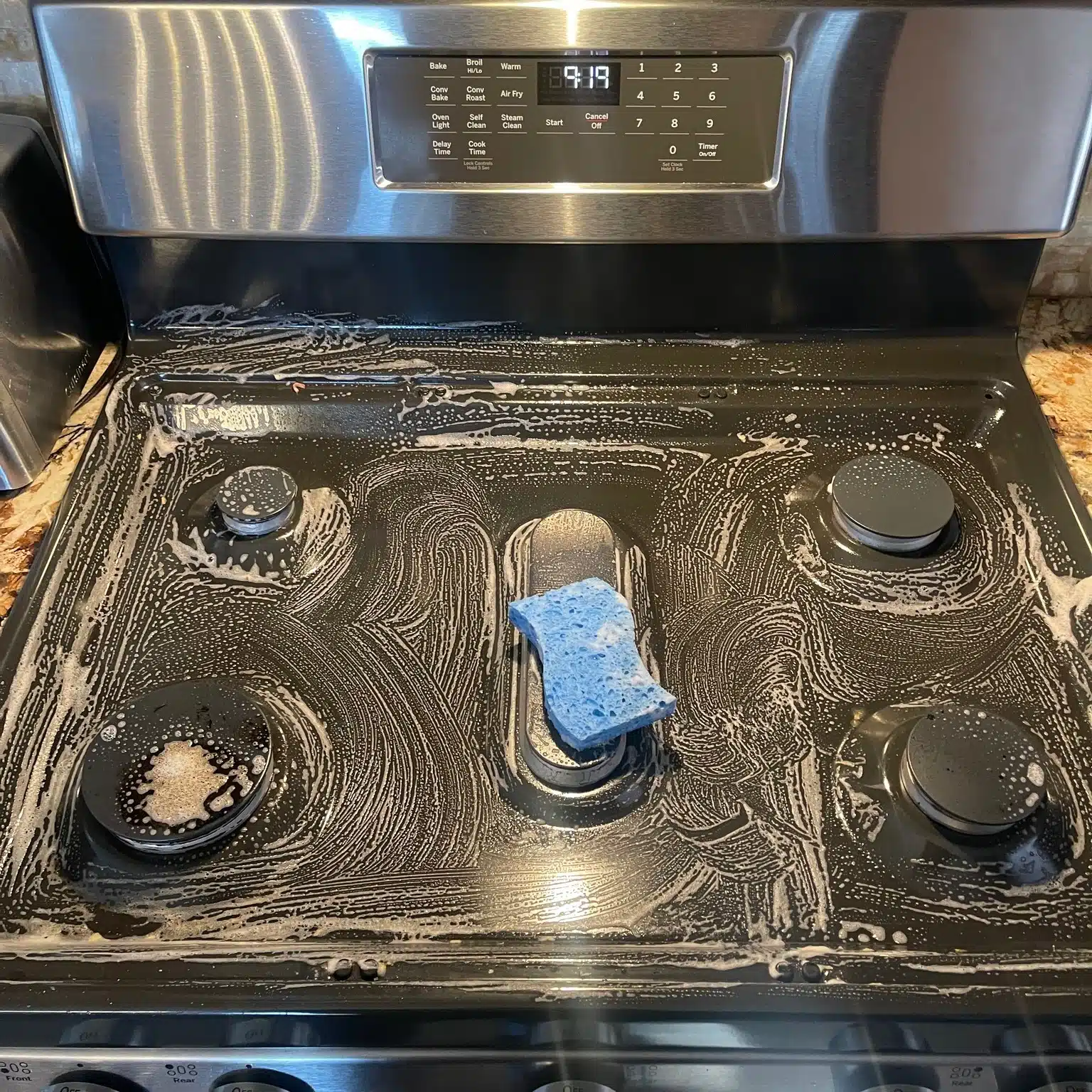 Professional stovetop cleaning in progress, showing a black gas stove covered with soap and cleaning solution as a blue sponge scrubs away grease, food residue, and buildup around the burners.