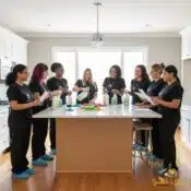 Professional cleaning team gathered in a bright kitchen for training, reviewing checklists, cleaning products, and microfiber cloths while wearing branded Sparkle & Shine uniforms and shoe covers.