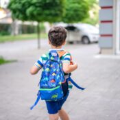 Back view of a child with a colorful dinosaur backpack walking on a sidewalk in a suburban area.