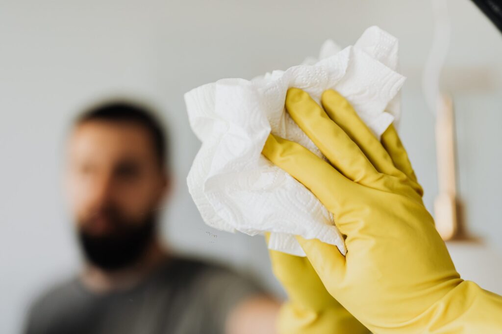 Blurred bearded man cleaning mirror with yellow glove and paper towel, emphasizing cleanliness.