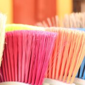 "Close-up of colorful broom bristles in a row — cream, pink, orange, and blue — fanned out against a blurred warm-toned background