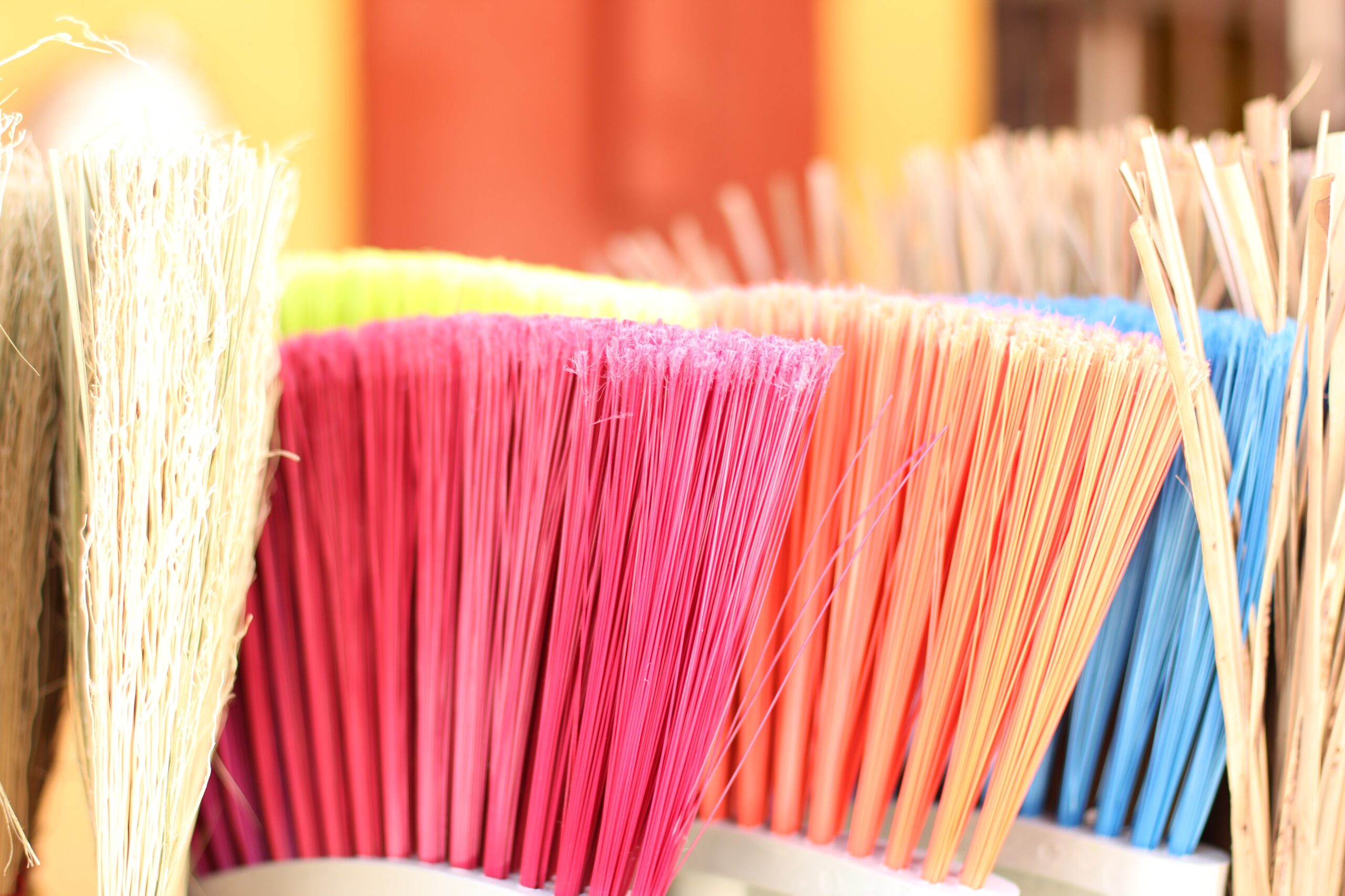 "Close-up of colorful broom bristles in a row — cream, pink, orange, and blue — fanned out against a blurred warm-toned background