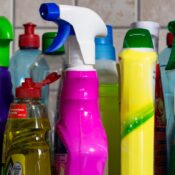 Row of colorful spray bottles and cleaning products lined up on a kitchen counter with a tiled backsplash.