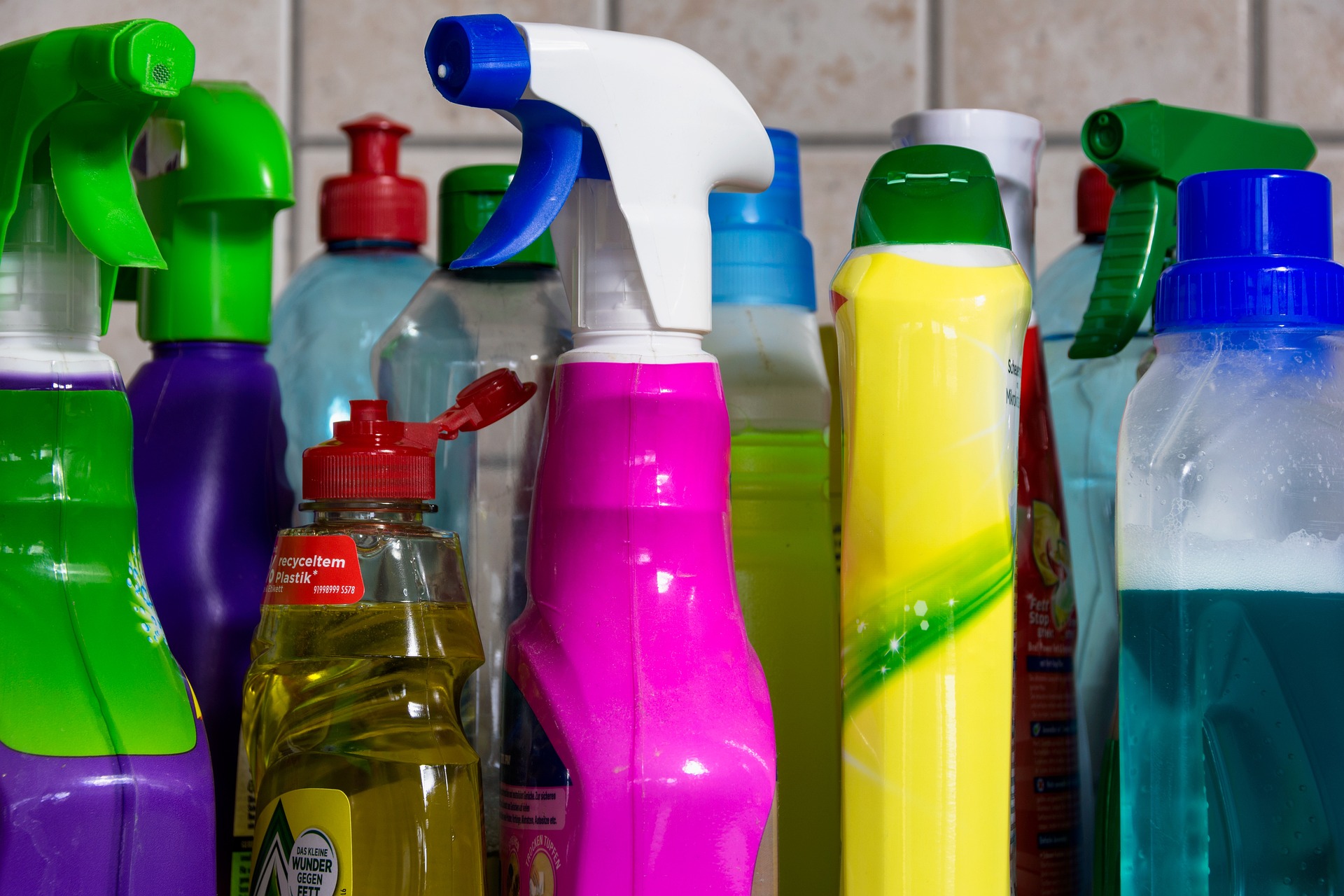 Row of colorful spray bottles and cleaning products lined up on a kitchen counter with a tiled backsplash.