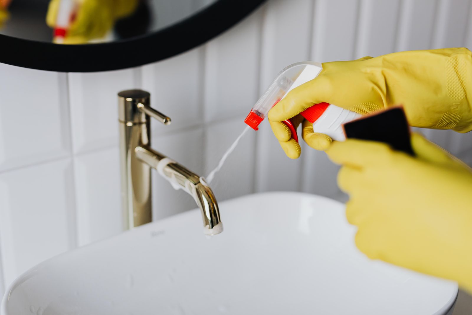 Gloved hands spraying and wiping a bathroom faucet during a deep cleaning